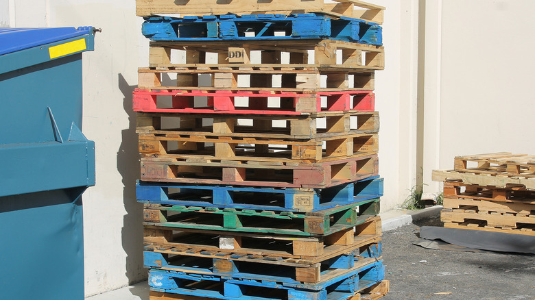 A stack of multi-colored pallets next to a dumpster against a white building