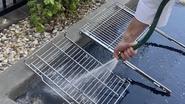 A hand and arm in a white sleeve sprays steel oven racks with water from a garden hose in a residential backyard.