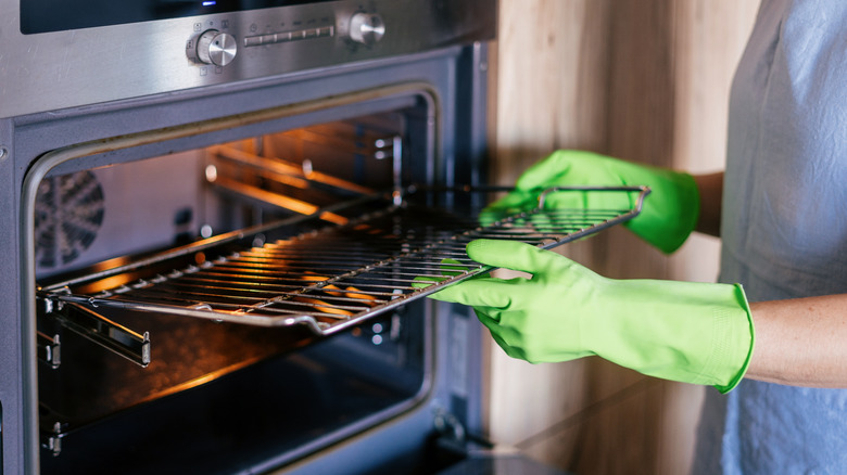 A woman wearing green gloves removes an oven rack from a wall oven.