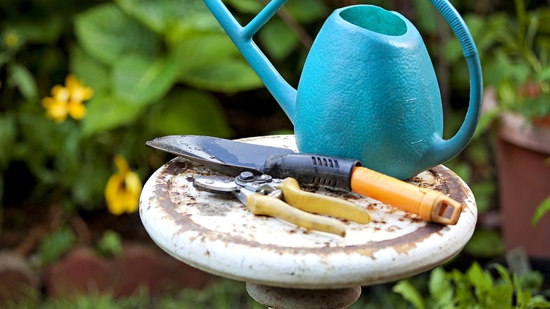 A hand trowel and pruners rest on a pedestal together with an aqua watering can in a garden