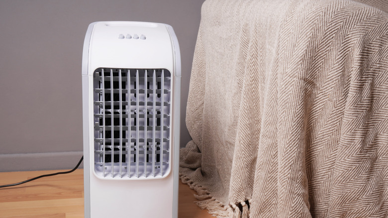 A portable AC unit next to a bed with a tan throw blanket in a bedroom.