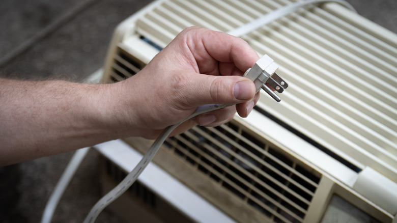 A person holds the plug of a white window AC unit in their hand.