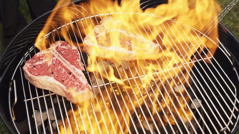 A pair of steaks cooking on a charcoal grill with flames leaping from it.