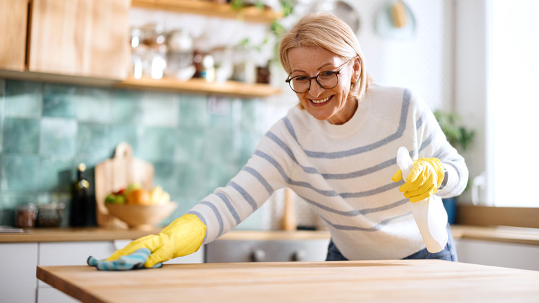 A smiling woman wearing yellow gloves cleans a kitchen countertop