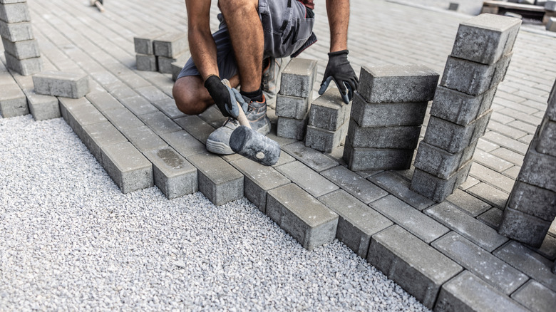 A man crouches while installing gray pavers on a compact base down a slight slope