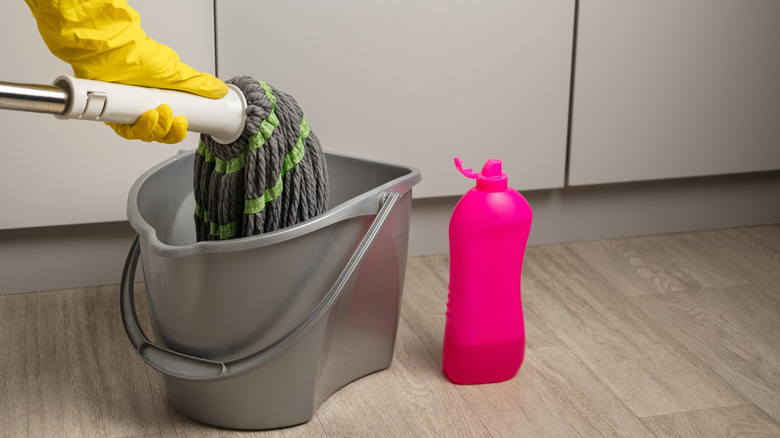A person dipping a mop in a bucket next to a pink bottle of soap