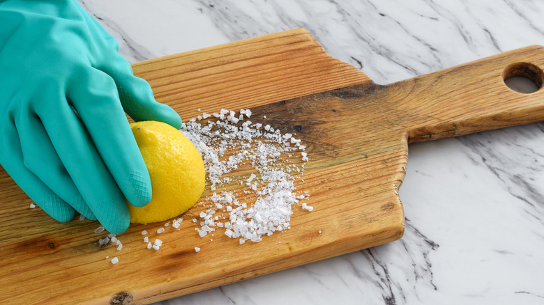 Closeup of person's hand in protective rubber glove scrubbing wooden cutting board with half lemon and coarse salt.