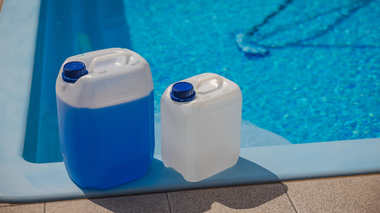 Two plastic chemical canisters with blue caps placed on the edge of a swimming pool, used for water treatment and maintenance in bright sunlight.