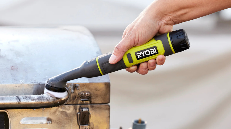 A person uses a cordless Ryobi detail scrubber to clean a dirty metal toolbox
