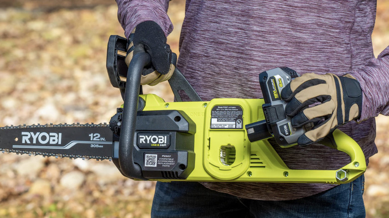 A man wearing long-sleeves and gloves holding a Ryobi Whisper Series chainsaw in front of his abdomen.