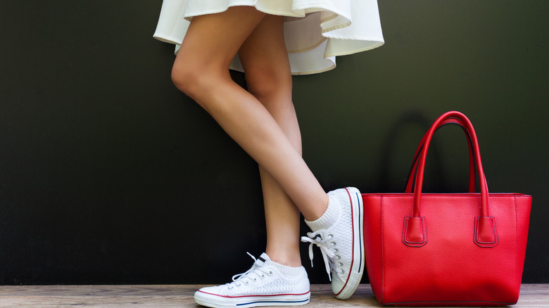 A fashionable woman in a dress sports bright white sneakers while a large red handbag rests nearby.