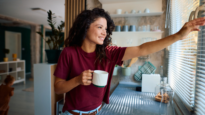 smiling young woman in kitchen holding coffee cup and opening window blinds