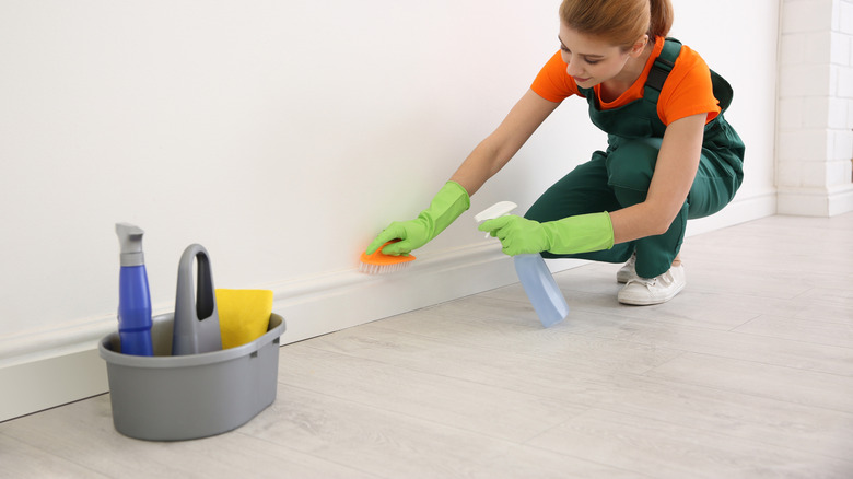 A person cleaning baseboards with spray cleaner and a scrub brush