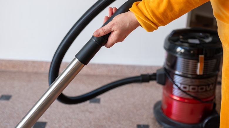 A woman in a yellow outfit vacuuming a tile floor with a vacuum attachment hose.