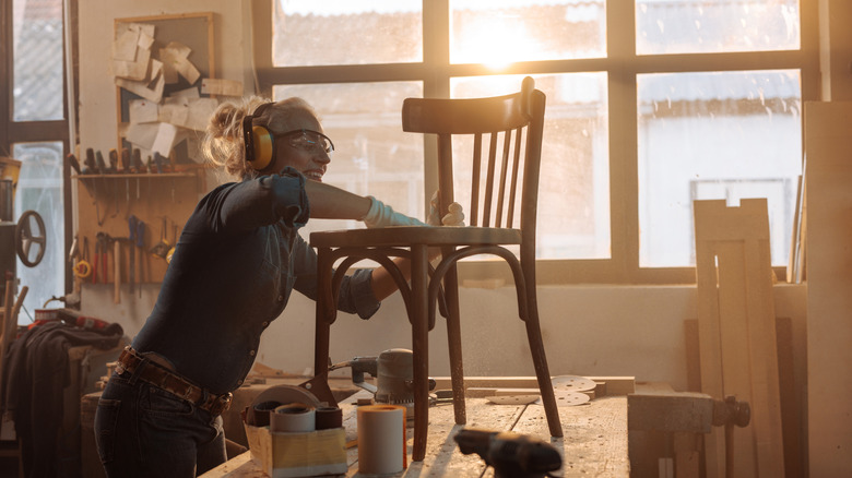 A woman smiles while sanding a chair in a light-filled workshop