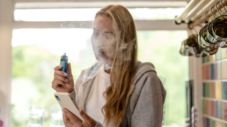 A young woman exhaling from a vape device while focused on her smartphone