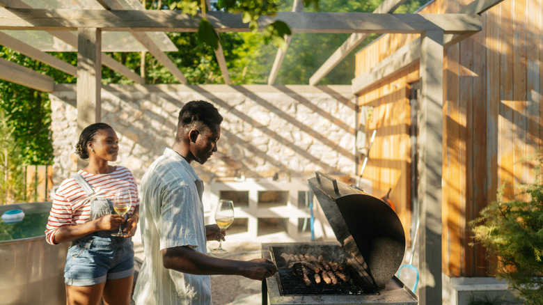 Person grilling meat on a grill outdoors with a woman drinking wine