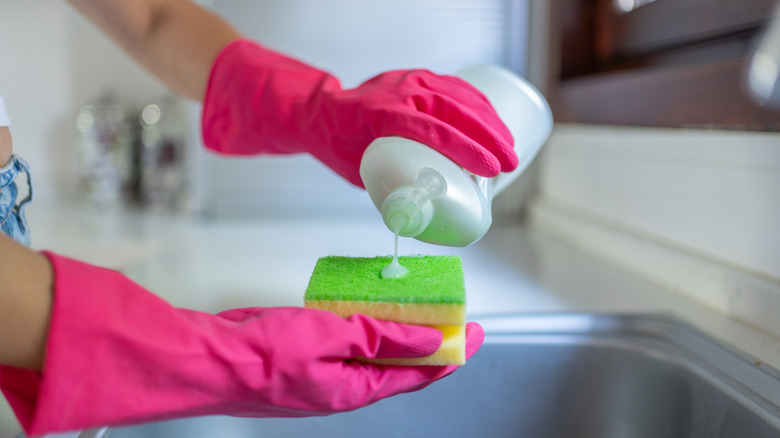 A person pouring dishwashing liquid onto a sponge over a sink