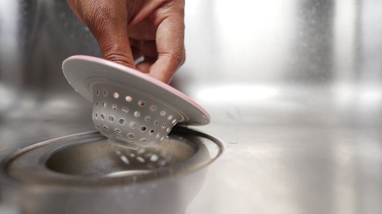 A hand lifts a plastic drain strainer from a kitchen sink drain hole.