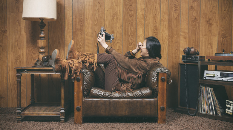 A retro-style woman holding a camera in one hand poses on a leather couch to take a selfie.
