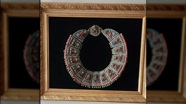 An intricate beaded necklace hangs against black velvet in an ornate frame.