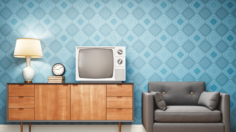 A retro-styled living room features a simple wooden TV stand that holds a TV, large white lamp, books, and a clock, next to a gray armchair.