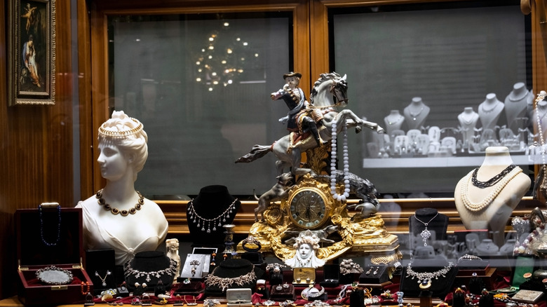 A jewelry shop display window with a faux Greek bust and an ornate gold clock amidst pieces of jewelry.
