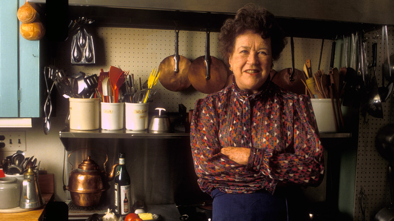 Julia Child standing in a kitchen with lots of utensils on the shelving unit behind her.