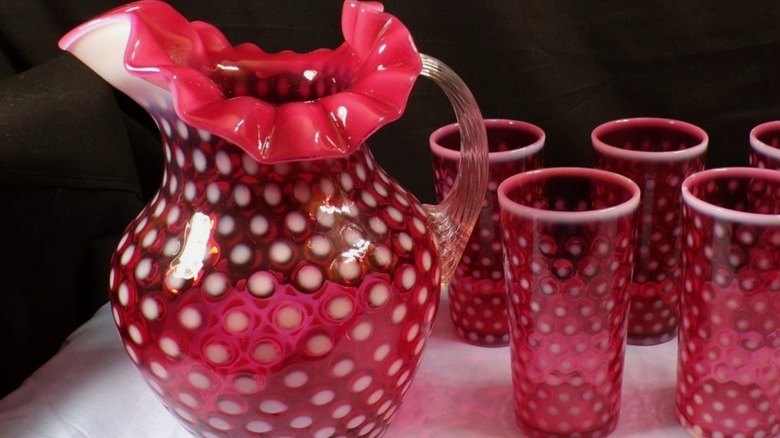 A red glass water pitcher with white polka dots next to drinking glasses with the same pattern.