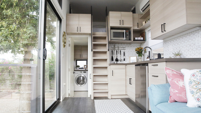 The interior of a tiny house with large glass windows, showcase the kitchen, part of the living room and loft bedroom.