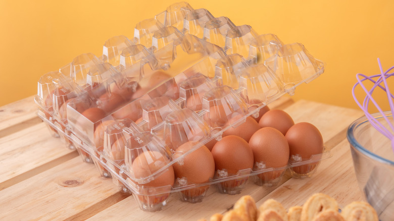 A clear plastic egg carton holds brown eggs and is resting on a countertop made of light wooden slats.