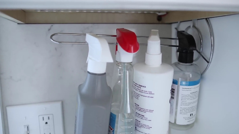 A wire paper towel holder mounted to the wall with cleaning supplies hanging on it.