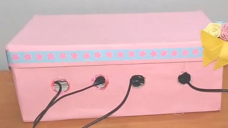 A shoe box is painted pink with holes cut out for cable wires