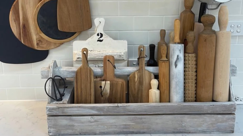 A collection of rolling pins and cutting boards is stored neatly on the kitchen countertop in an old wooden box