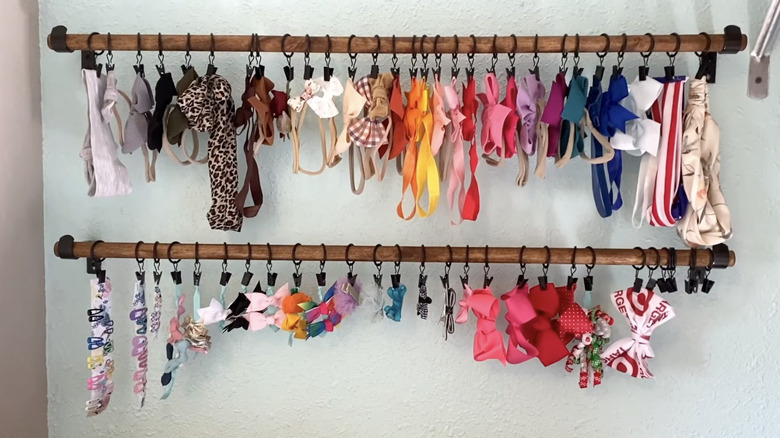 Two parallel wooden curtain rods have curtain rings holding hair bows and clips in a variety of colors