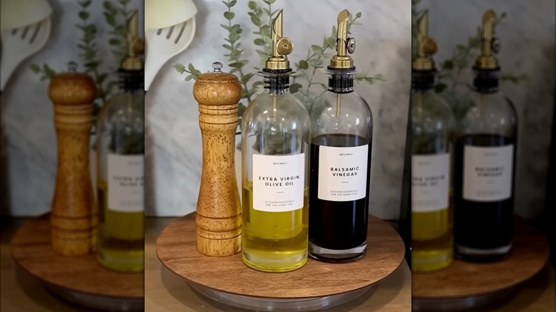Bottles of oil and vinegar, a salt grinder, and a potted plant sit on a wooden lazy Susan on a kitchen counter.
