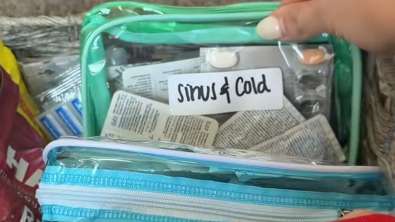 A hand holds a green-rimmed transparent plastic toiletry bag with various medicines inside and a label that says sinus and cold.