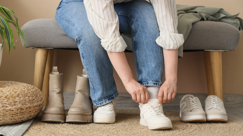 A person leaning over to pull on white shoes while sitting on a bench near a home entry.