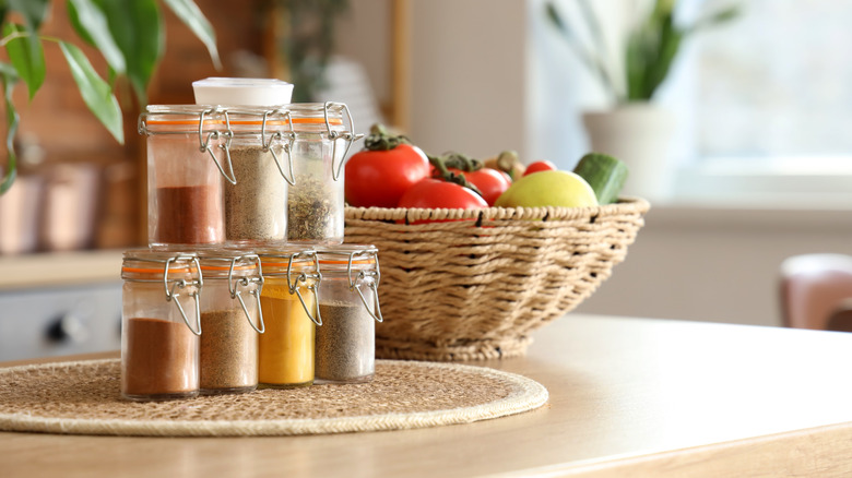 Spices in sealed jars stacked on a woven mat on a kitchen countertop nearby a bowl of fruit and vegetables.