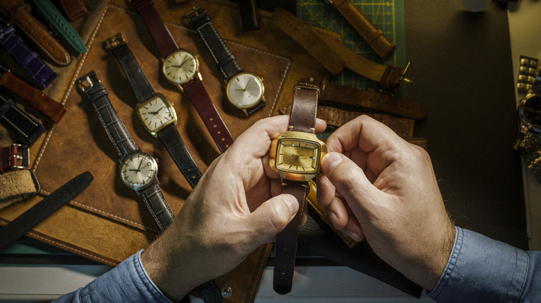 Many watches laid out on a desk top with a pair of hands holding one watch and winding it.