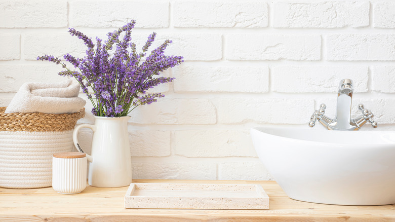 A bathroom countertop with lavender in a vase, a small tray, round pot, and a towel in a basket