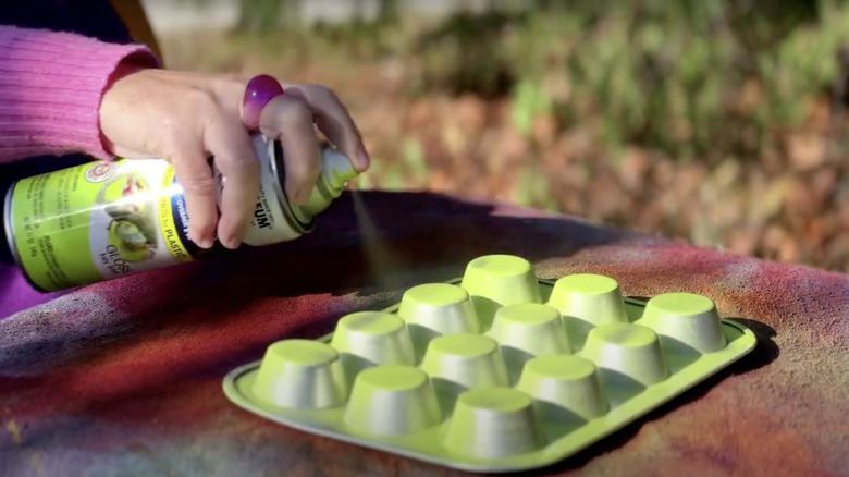 A woman paints a muffin tin outside using highlighter yellow spray paint.