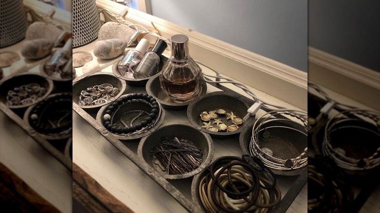 An antique metal cupcake pan sits on a bathroom countertop, each cup filled with a different type of beauty product or accessory.