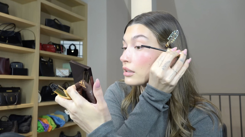 Hailey Bieber applying makeup with shelves of handbags in the background