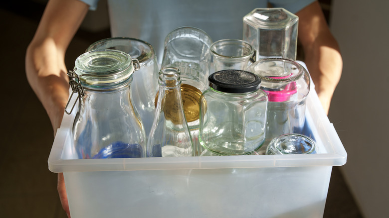 A person holding a plastic bin full of empty glass jars of different shapes and sizes