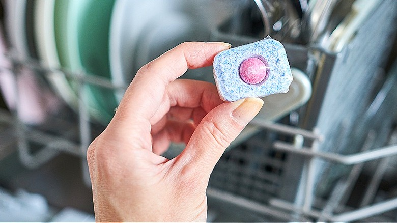 Close-up of a hand holding a dishwasher pod or tablet with blurred dishes in a dishwasher in the background