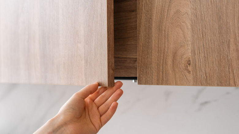 Cropped shot of woman hand is opening a wooden cabinet door in contemporary kitchen, highlighted by natural lighting