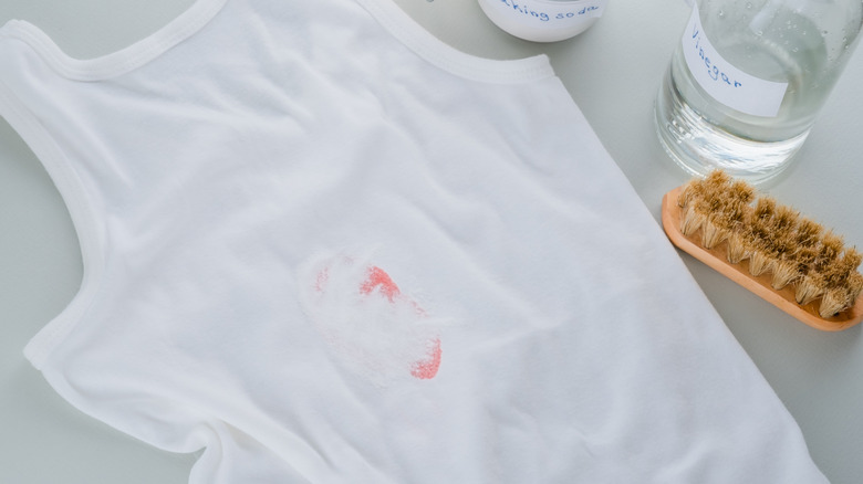 A white tank top with a red stain beside a bottle of white vinegar, a coarse brush, and a jar of baking soda.