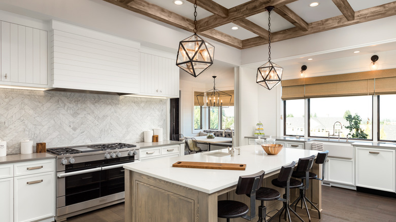 A modern kitchen with architectural wood beams in the ceiling, recessed lights, wood cabinets, tall herringbone backsplash, and soffits with recessed lights
