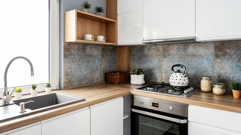 Small white kitchen with stylish tiles, wooden cabinets and sink with faucet near the window.